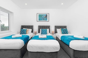 A hotel room with three beds, each with a grey headboard, white bedding, and blue accents. There is a framed abstract painting on the wall above the beds. - 5 Bed House - Parking - Near Birmingham Centre (Birmingham)