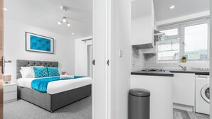 Un dormitorio moderno con una cama tapizada en gris, una manta azulada y una obra enmarcada en la pared. Junto a ello, una cocina compacta con lavadora y una ventana pequeña.