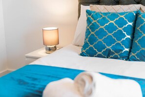 Een net opgemaakt bed met een blauwe deken, een bedlampje en decoratieve kussens.