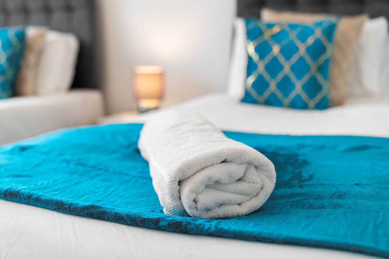 A neatly made bed with a rolled white towel, a blue textured blanket, and a geometric patterned pillow.