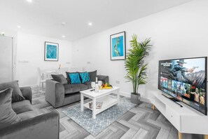 A modern living room with a grey sofa, a white coffee table, and a flat-screen TV displaying a movie.