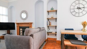 Ein Wohnzimmer mit grauem Sofa, einem Holzesstisch mit Stühlen, einem Kamin, einem Bücherregal und einer Wanduhr.