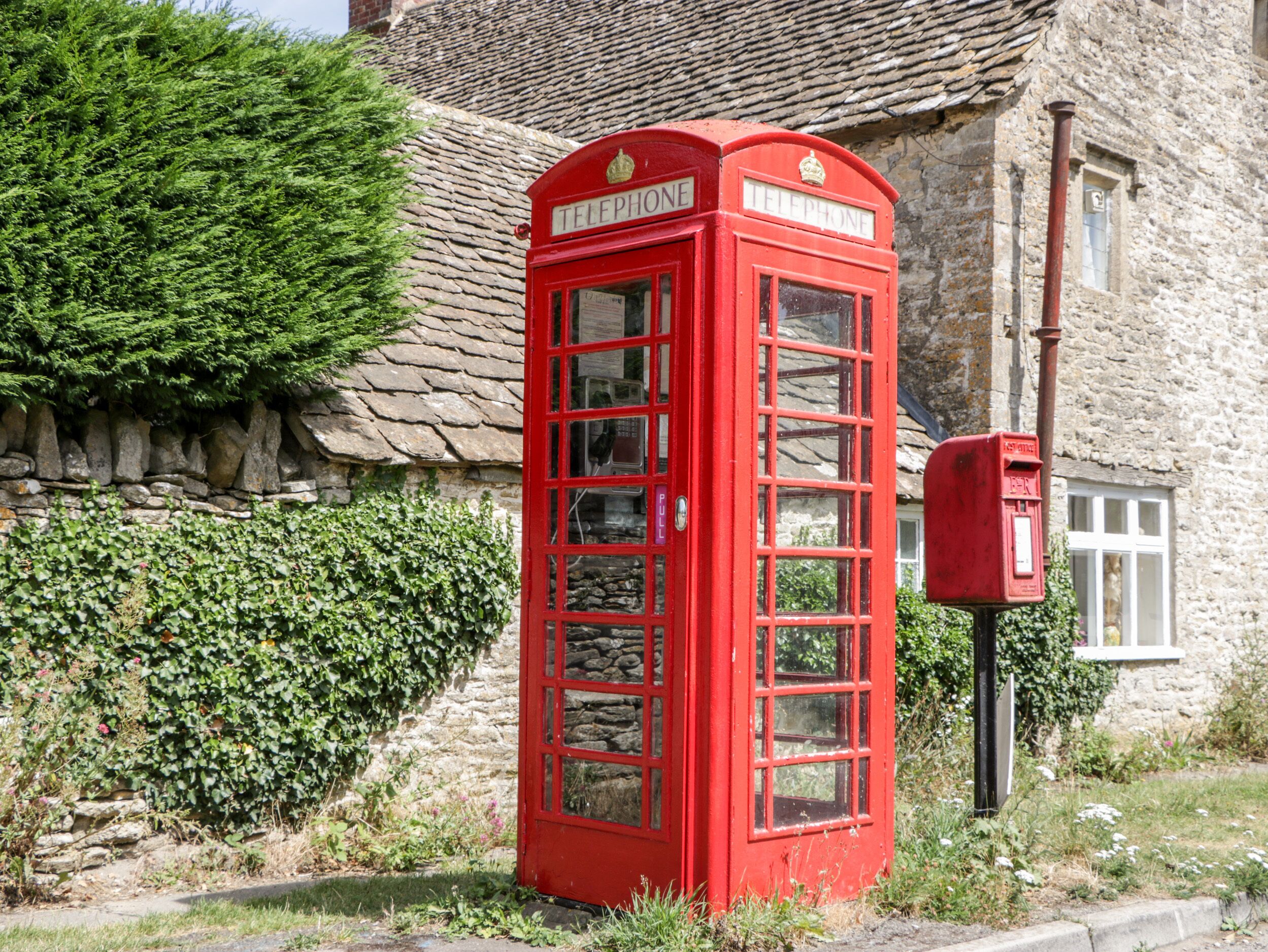 A red British telephone booth with a stone wall and a mailbox in front.