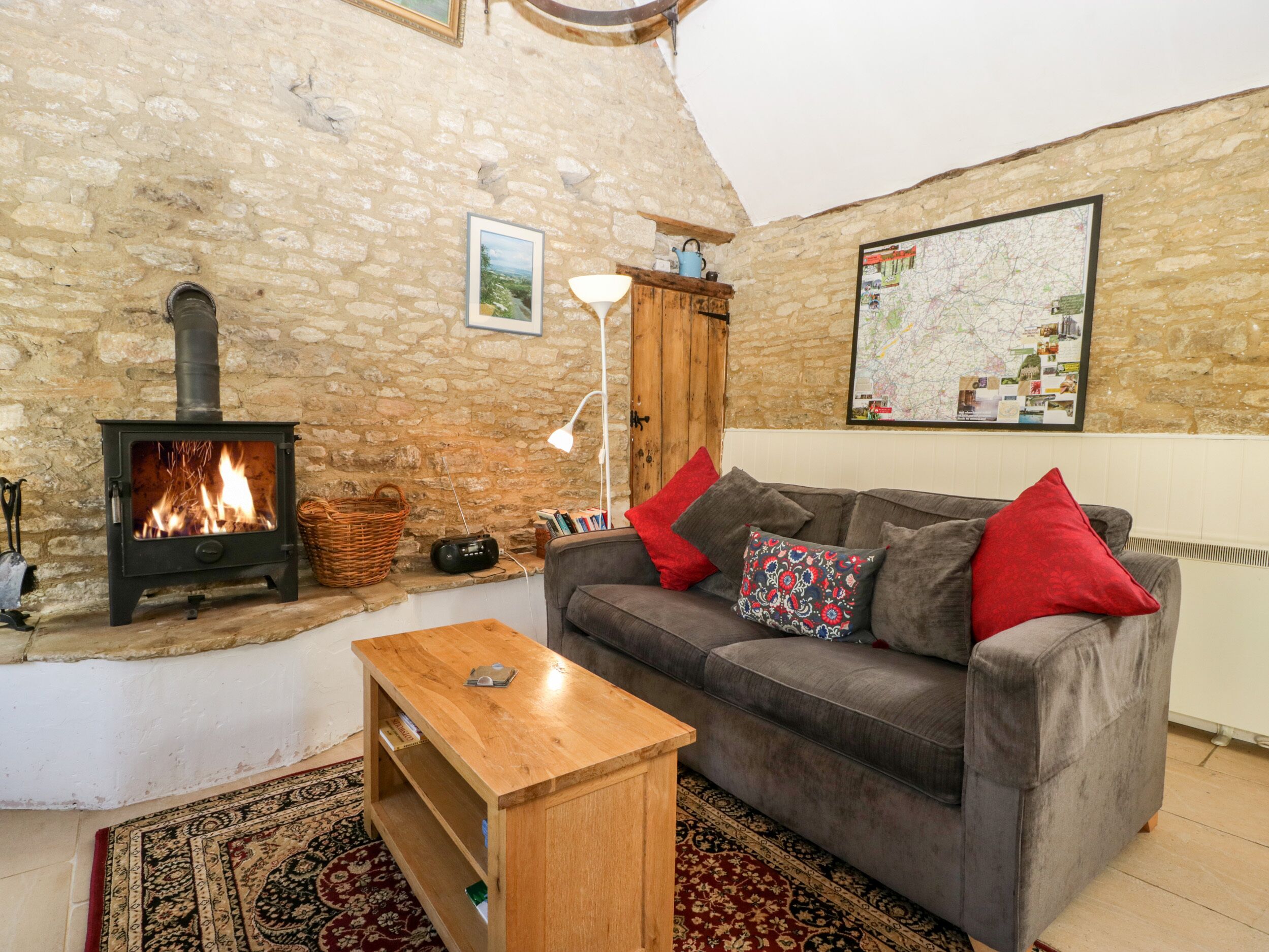 A cozy living room with a stone wall, a fireplace, a sofa with red cushions, a wooden coffee table, and a map on the wall.