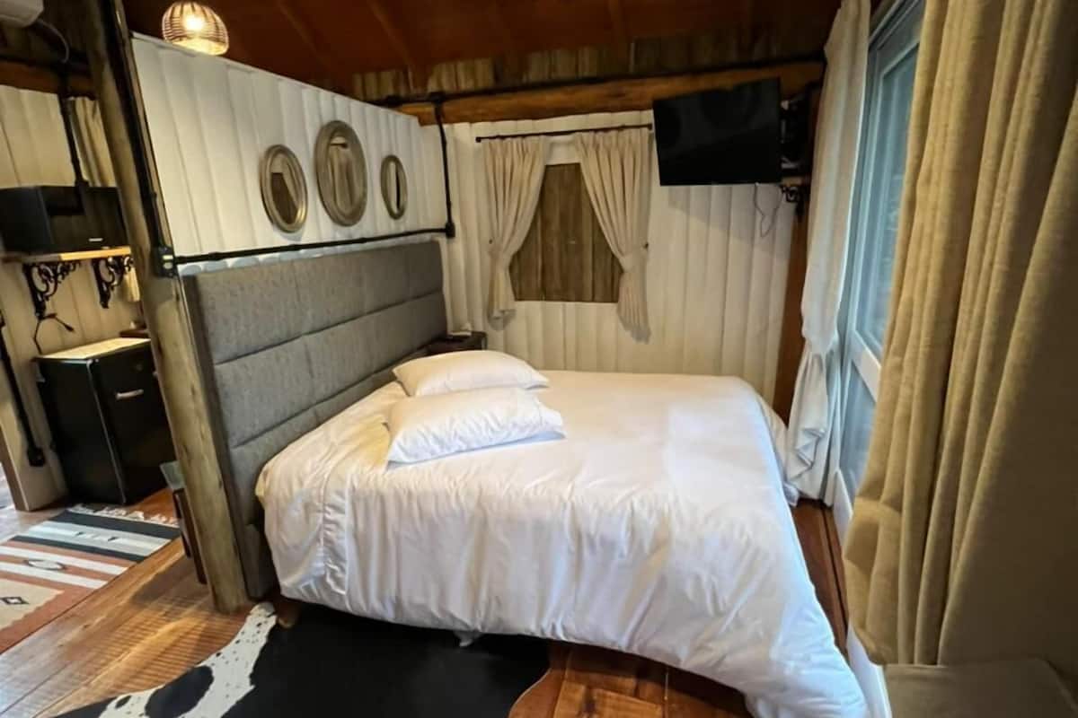 A cozy room with a bed, a wooden ceiling, a window with curtains, and a mounted TV.