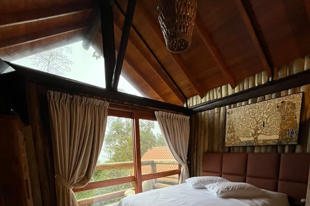 A cozy bedroom with a large bed, wooden ceiling beams, a window with curtains, and a colorful striped rug.