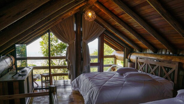A cozy attic bedroom with a wooden ceiling, a large bed, a desk, and a view of greenery outside. - Pousada Parador 33 (Biguacu)