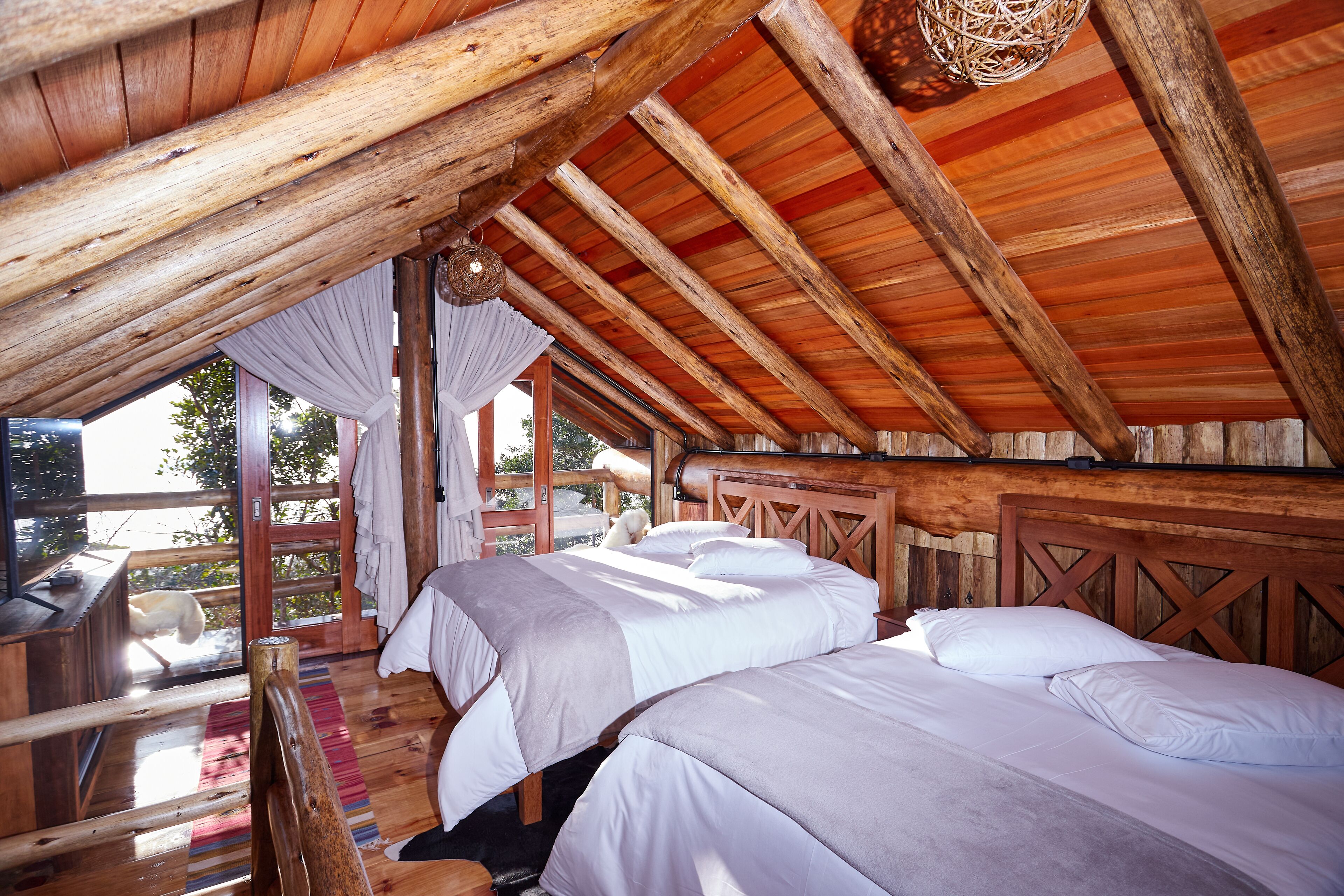 A rustic cabin with wooden beams, a loft-style bedroom with two beds, a large window, and a view of the outdoors.