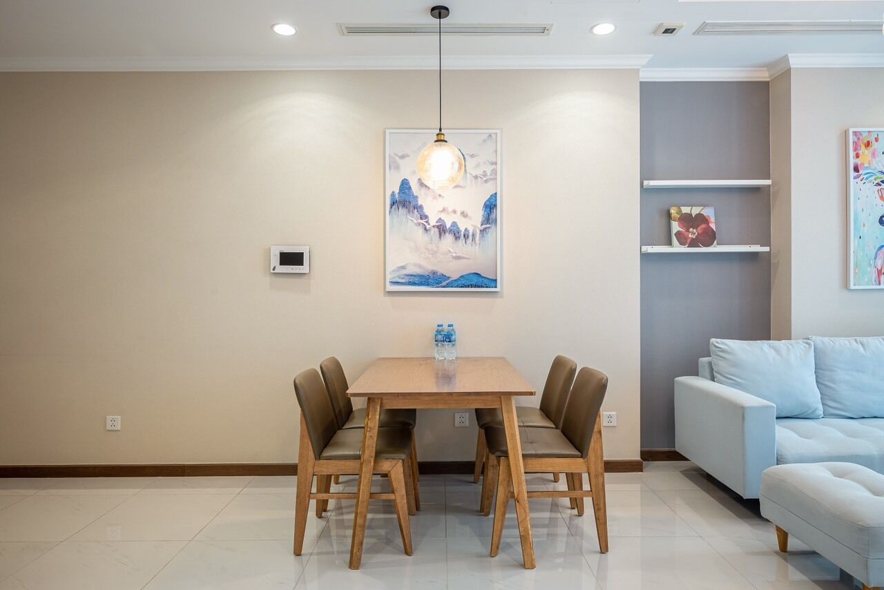 Un comedor con una mesa de madera y cuatro sillas, una lámpara colgante, un termostato en la pared y un sofá al fondo.
