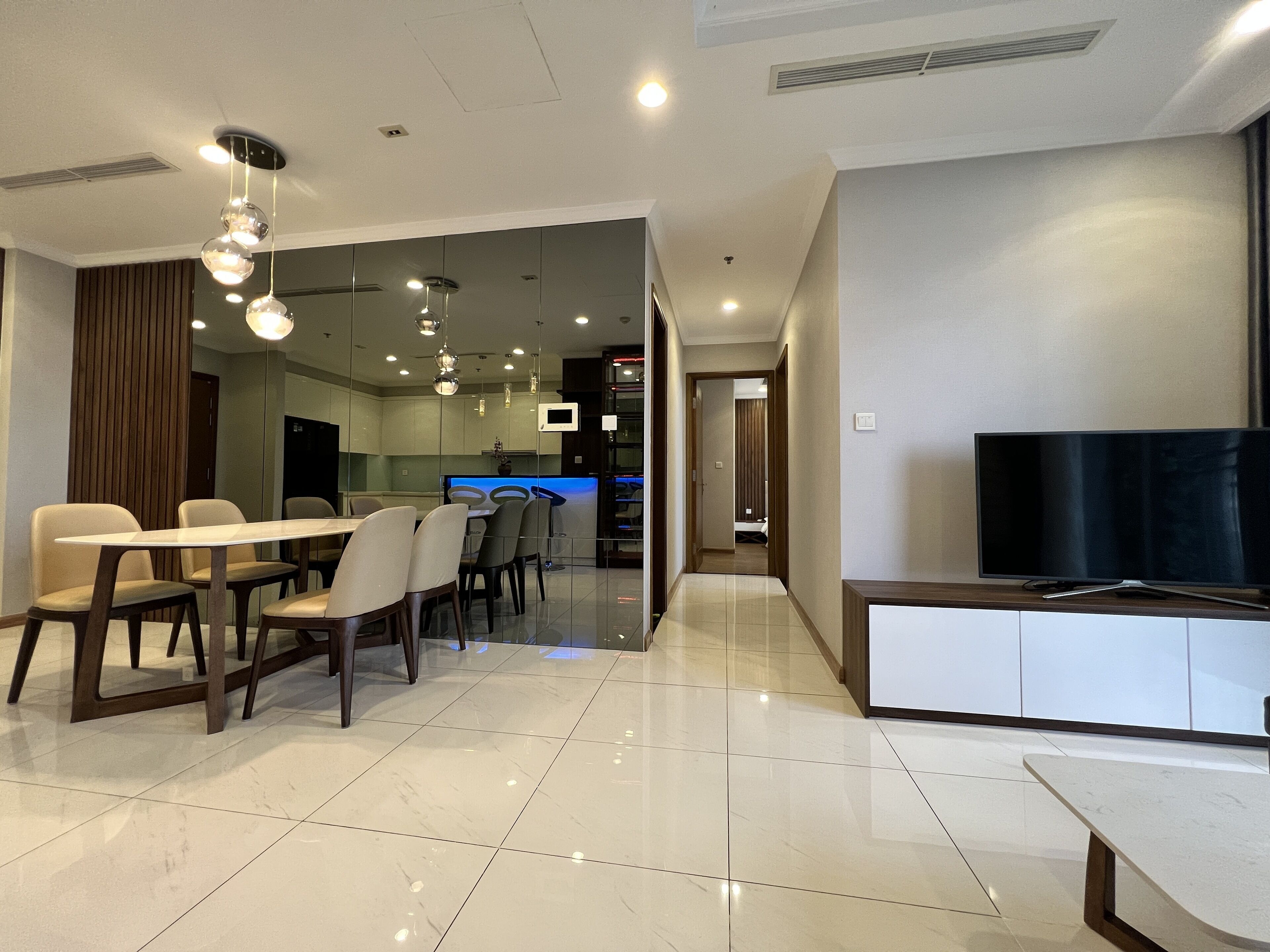 A modern living room with a dining area, a flat-screen TV, and a kitchen visible through a glass partition.