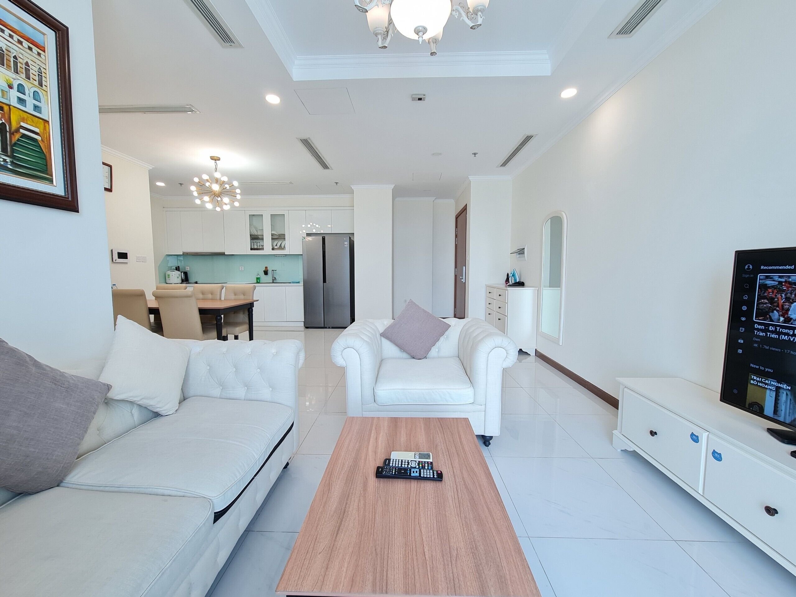 Ein modernes Wohnzimmer mit Sofa, Sessel und Couchtisch. Im Hintergrund ist eine Küche mit Kühlschrank und Schränken zu sehen.