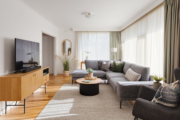 A modern living room with a flat-screen TV, a wooden cabinet, a grey sofa set, a round coffee table, and a large window with curtains.