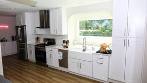 A modern kitchen with white cabinets, stainless steel appliances, and a window with a view of greenery. - The Euclid 1925 Bungalow Located in Downtown (St. Petersburg)