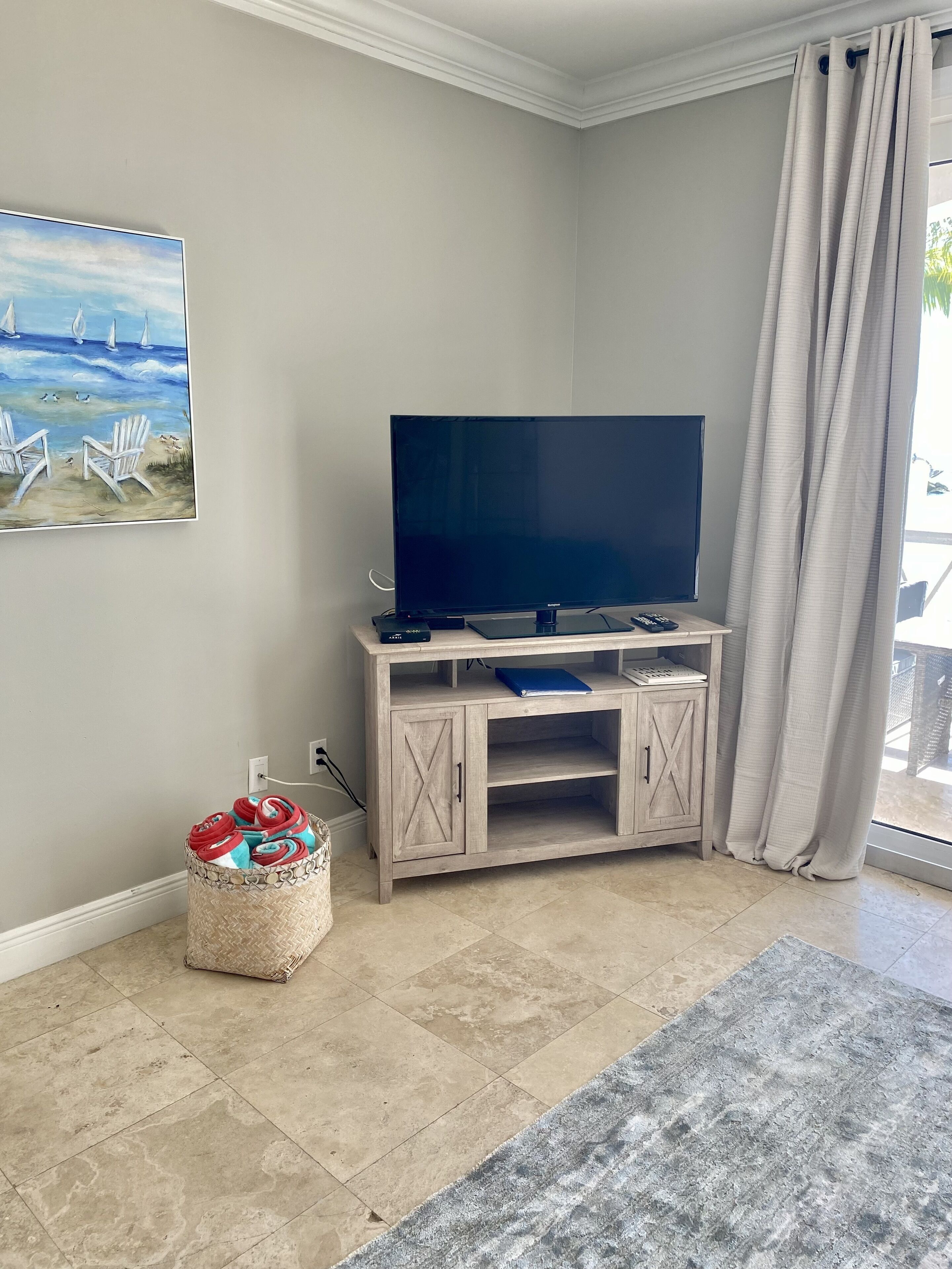 Un salon avec un téléviseur à écran plat posé sur un meuble télé en bois, un tableau représentant des voiliers accroché au mur, un panier en osier contenant des objets rouges et bleus, et un tapis gris.