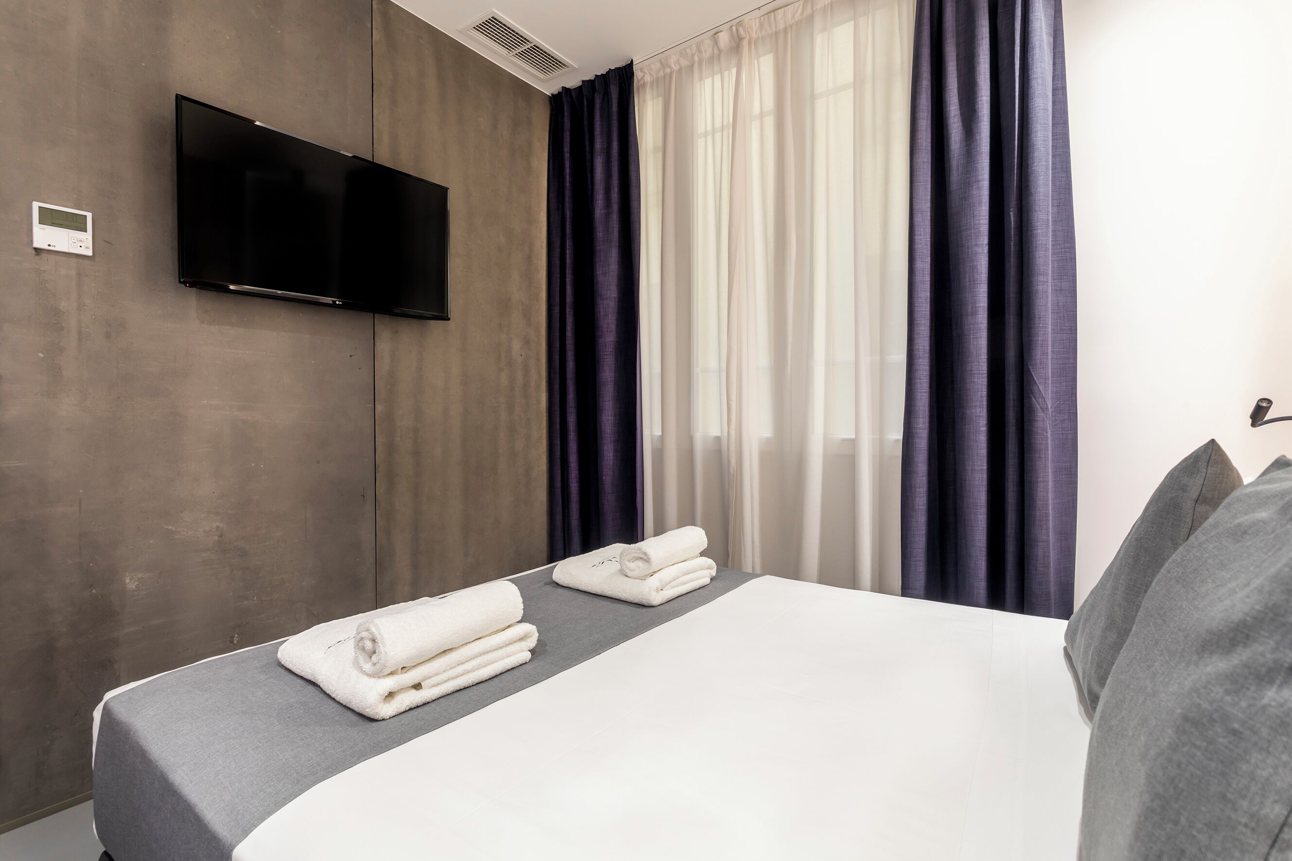 A hotel room with a bed, a flat-screen TV mounted on the wall, and a window with curtains.