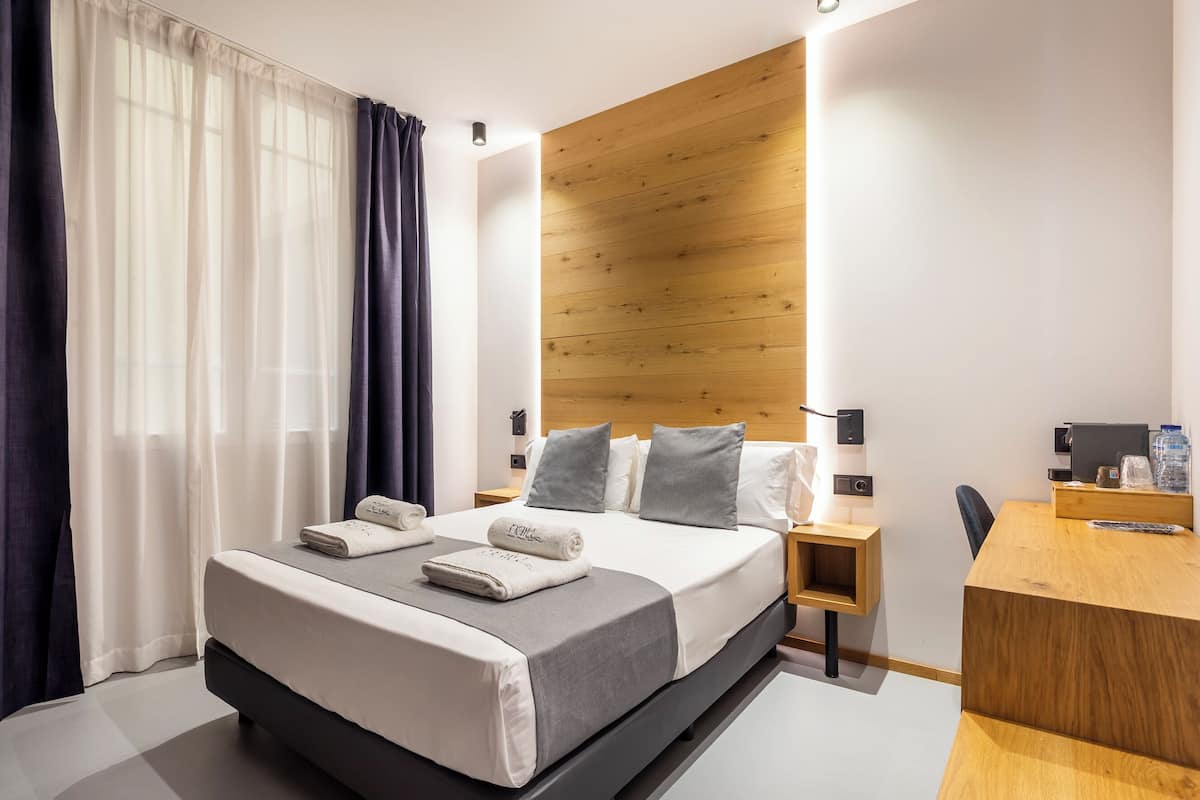 A modern hotel room with a large bed, a wooden headboard, a desk, and a chair.