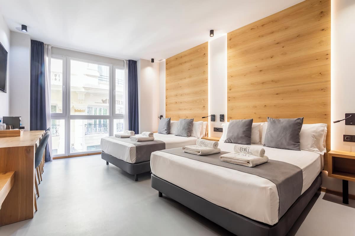 A modern hotel room with two beds, a wooden accent wall, a large window, and a desk.