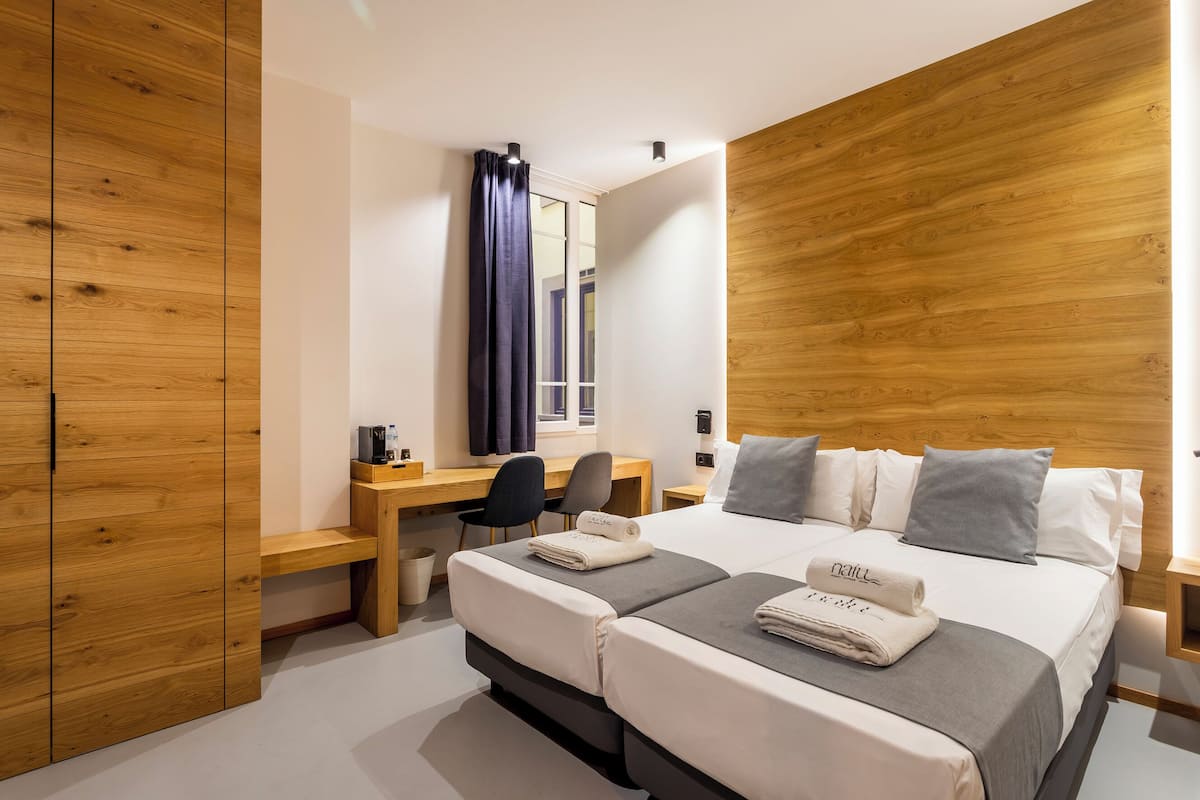 A modern hotel room with a wooden accent wall, a desk with a chair, a bed with two pillows, and a bedside table.