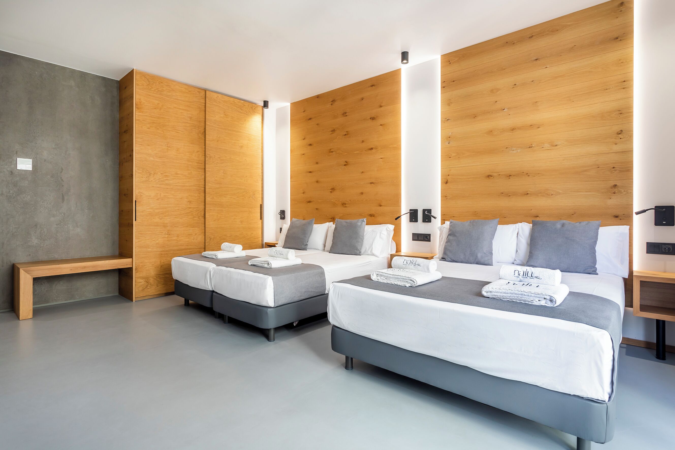 A modern hotel room with two beds, a wooden wardrobe, a bench, and a grey floor.