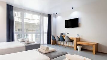 Habitación de hotel con dos camas, un escritorio con sillas, un televisor y vistas a edificios a través de amplias ventanas.