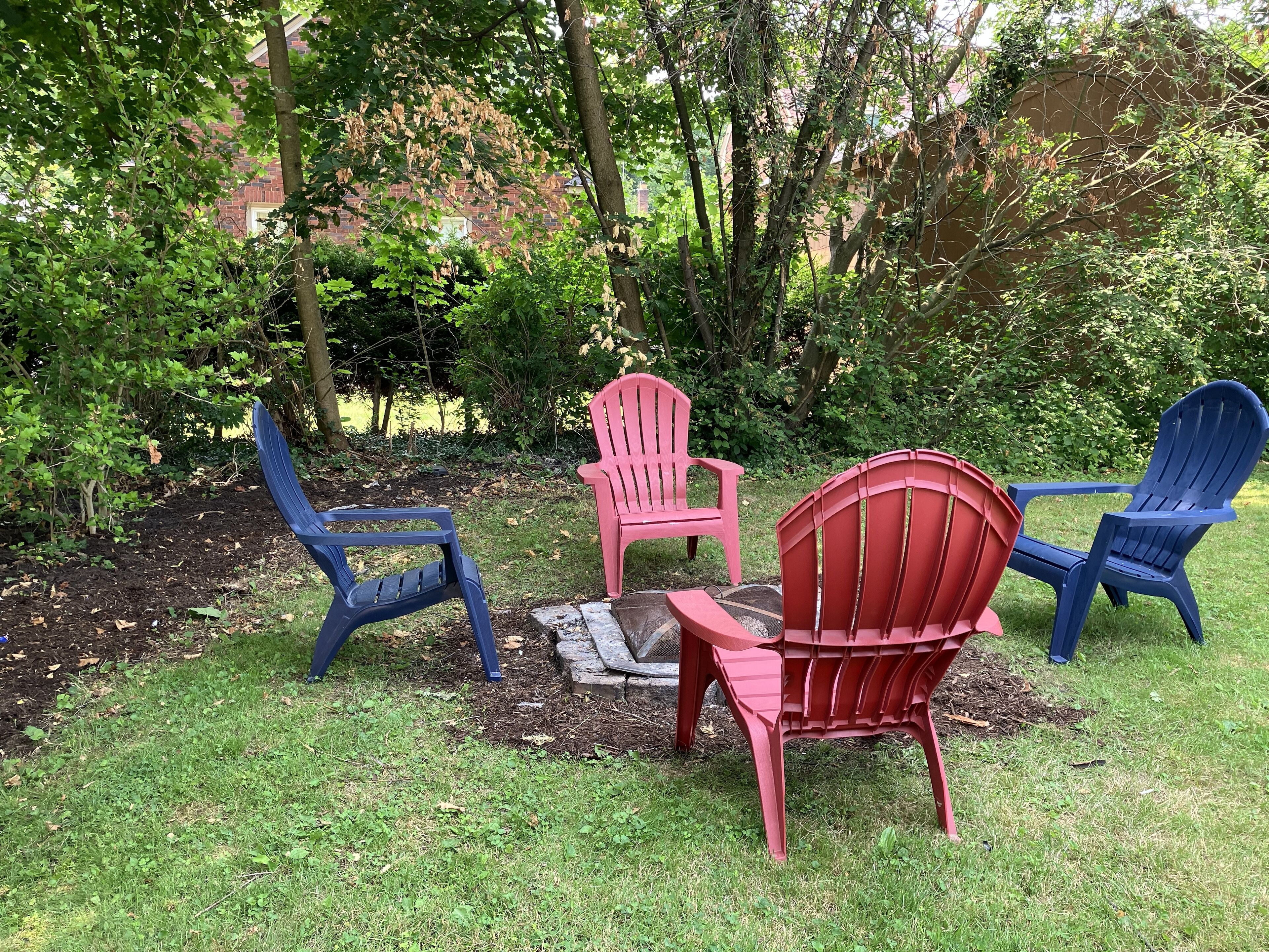 Ein Garten mit vier Adirondack-Stühlen in Blau, Rot und Rosa, die um eine Feuerstelle angeordnet sind.
