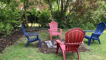 Ein Garten mit vier Adirondack-Stühlen in Blau, Rot und Rosa, die um eine Feuerstelle angeordnet sind.
