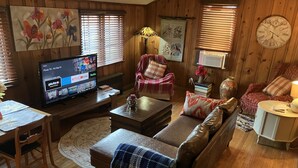 Un salón acogedor con suelo de madera, un televisor mostrando aplicaciones de streaming, un sofá, una silla y una pequeña mesa.