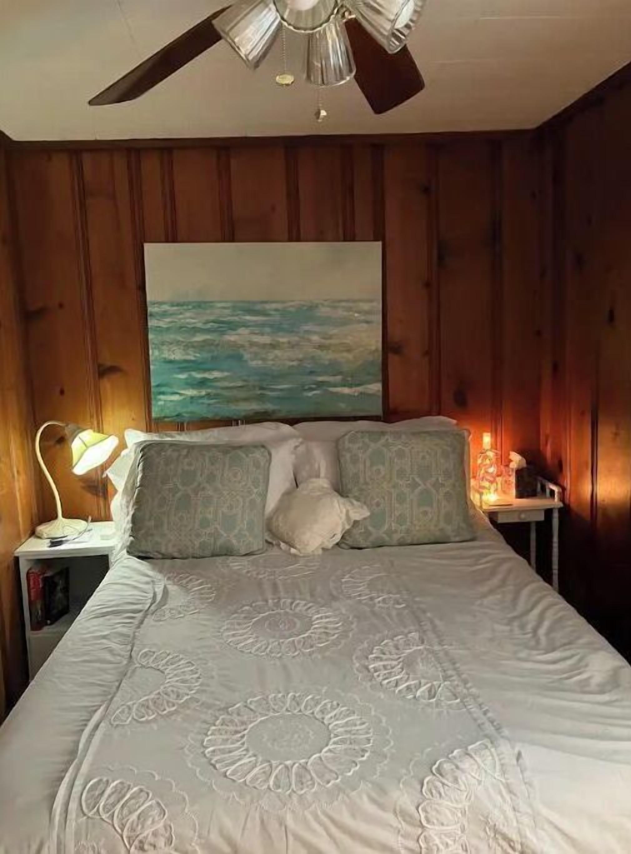 Et soveværelse med en væg i træpaneler, en ventilator i loftet, et maleri af et sølandskab, en seng med mønstrede puder og et natbord med en lampe.