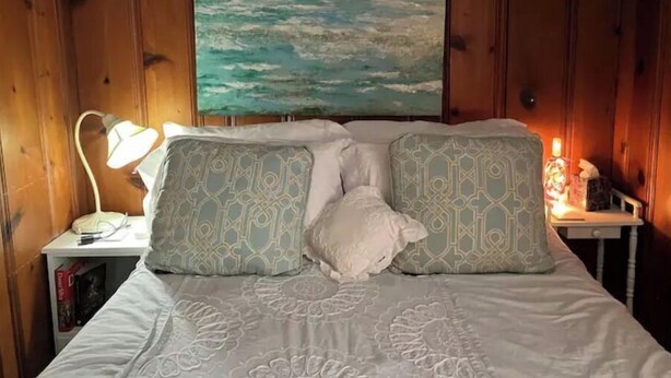 Un dormitorio con cama, almohadas estampadas, un cuadro del océano, una mesita de noche con lámpara y una pequeña mesa con lámpara.