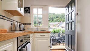 A compact kitchen with white cabinets, a wooden countertop, and a view of the garden through an open door. - Wood Cottage (Lynton)