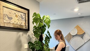 A person adjusting a large potted plant on a wooden table in a room with a gray wall, a framed artwork, and a ceiling vent.