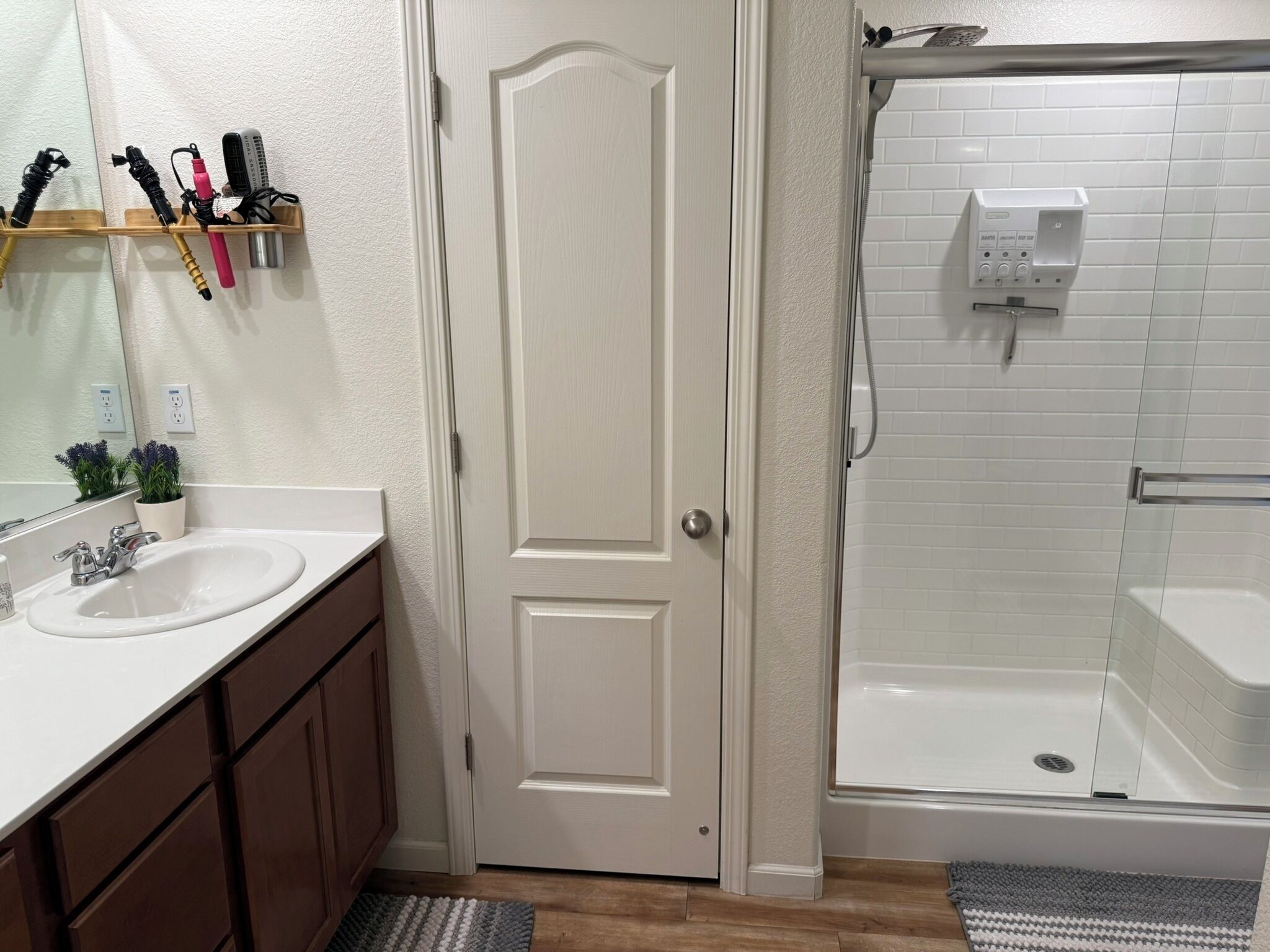 Ein Badezimmer mit einem weißen Waschbecken, einem Spiegel und einer Dusche. Eine Tür mit Griff ist vorhanden, sowie ein Ablagefach mit Toilettenartikeln.