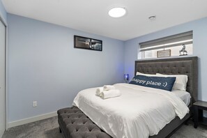 A bedroom with a bed, a headboard, a bedside table, a bench, a wall-mounted picture, and a window with blinds.