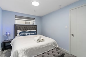 A bedroom with a tufted headboard, a bed with a white comforter, a nightstand with a lamp, and a door to the right. - Modern, cozy and private suite basement in South West Edmonton (Edmonton)