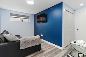 Ein Zimmer mit einem an einer blauen Wand befestigten Fernseher, einem schwarzen Sofa mit Decke, einem kleinen Tisch mit Gegenständen und einer Tür.