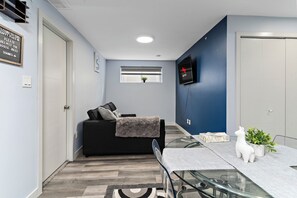 Ein zeitgemäßer Wohnraum mit einer Couch, einem Glastisch, einem Fernseher und einem Fenster mit Jalousien.