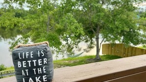 Un cojín con el texto "LIFE IS BETTER AT THE LAKE" sobre una barandilla de madera.