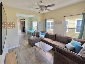 Ein Wohnzimmer mit einem braunen Ecksofa, einem kleinen weißen Tisch und einem Deckenventilator. Im Hintergrund ist ein Essbereich mit Tisch und Stühlen zu sehen.