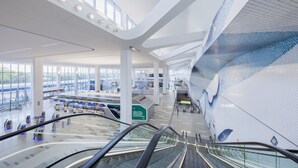 A modern airport terminal with a curved ceiling, escalators, and a large mural.