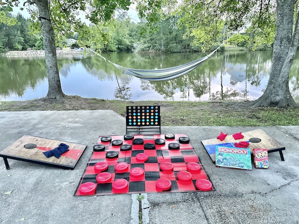 Un jeu de dames est installé sur une surface en béton, près d’une étendue d’eau. On peut apercevoir un hamac et un jeu de Monopoly à proximité.