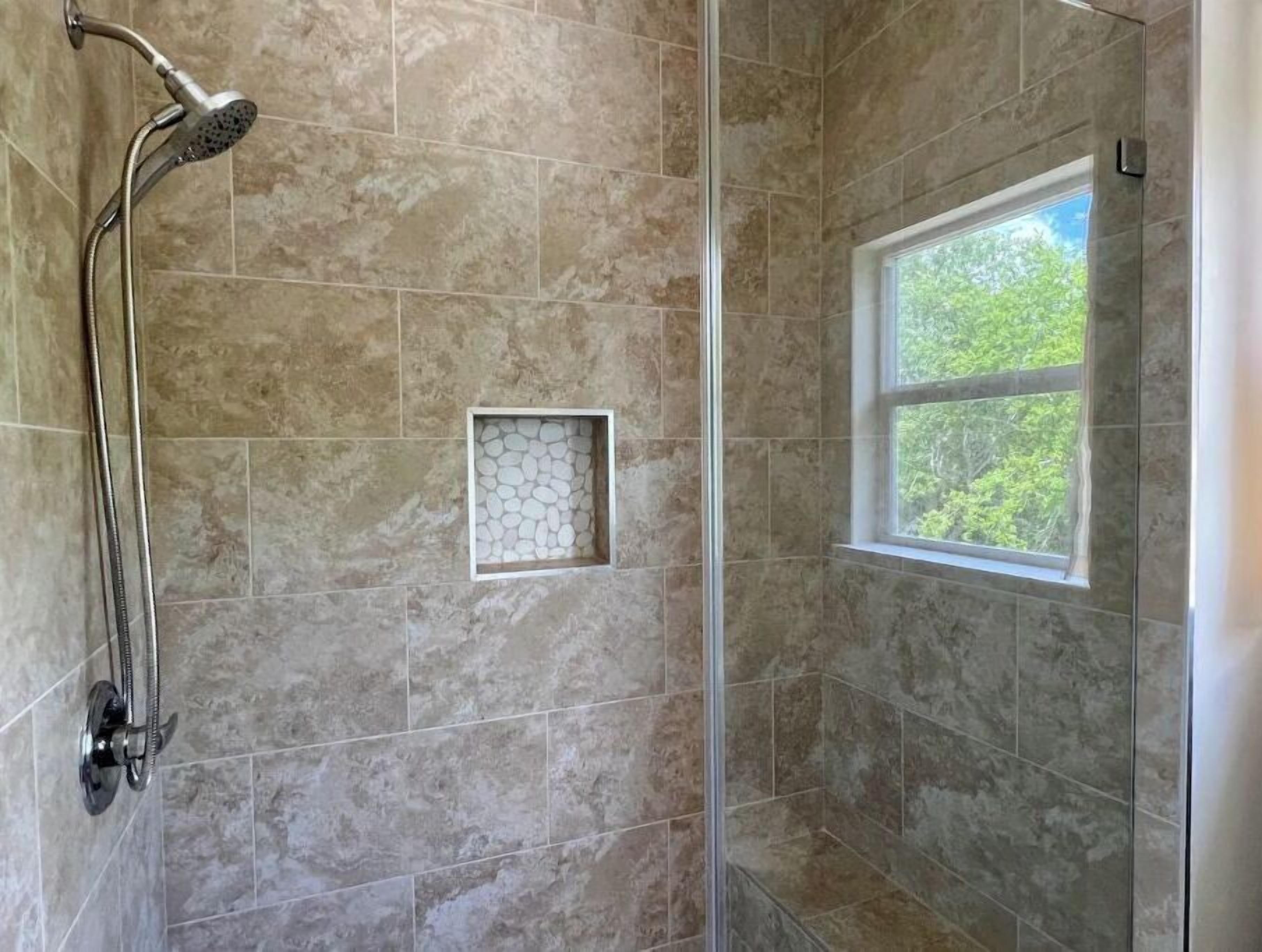 Una ducha con puerta de vidrio, una ventana con vista a árboles y un pequeño detalle cuadrado de azulejos en la pared.