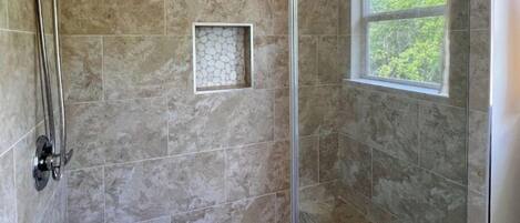 Una ducha con puerta de vidrio, una ventana con vista a árboles y un pequeño detalle cuadrado de azulejos en la pared.