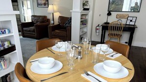 Mesa de comedor preparada para cuatro, con platos blancos, fuentes y vasos transparentes, rodeada de sillas de cuero marrón.