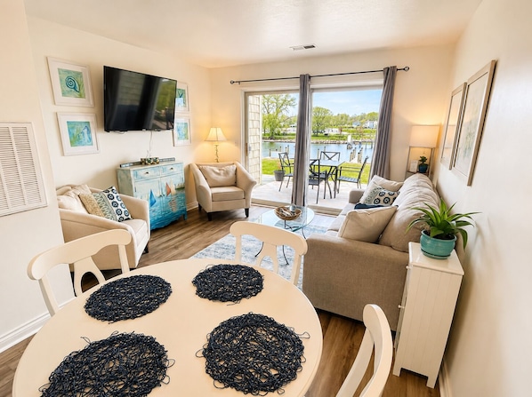 A modern living room with a TV, sofa, chairs, a dining table, and a view of a garden and pool. - Riverfront Retreat! Walk to Downtown/Near Beaches! (South Haven)