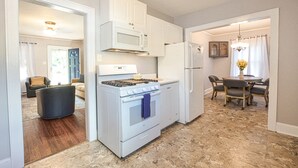 A modern kitchen with white cabinetry, a stove, microwave, and refrigerator. There is a dining area with a table and chairs. - Cozy Cottage minutes to Downtown (Winston-Salem)