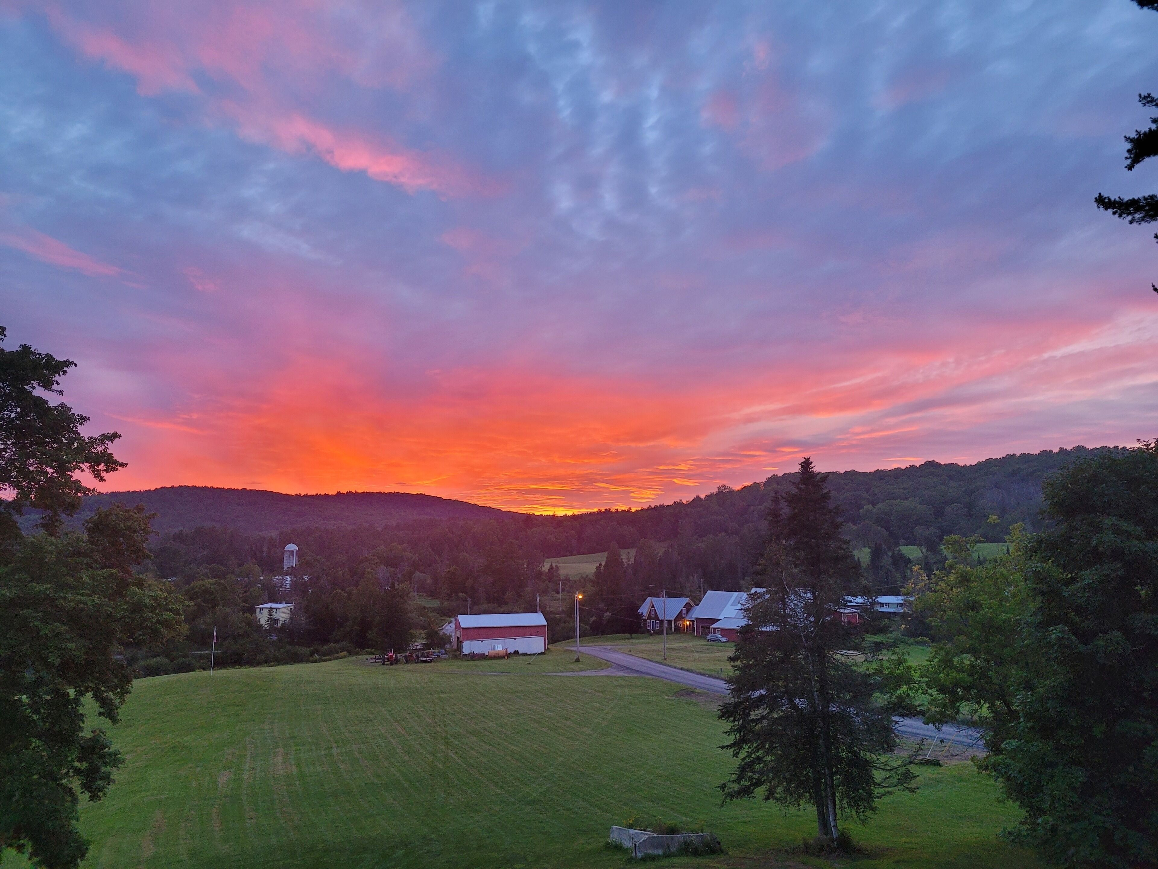 A rural landscape with a sunset, featuring a barn, a farmhouse, and rolling hills.