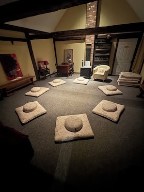 Een kamer met een cirkelvormige opstelling van vierkante kussens op de vloer, een open haard, een eettafel met stoelen, een boekenkast en een deur.