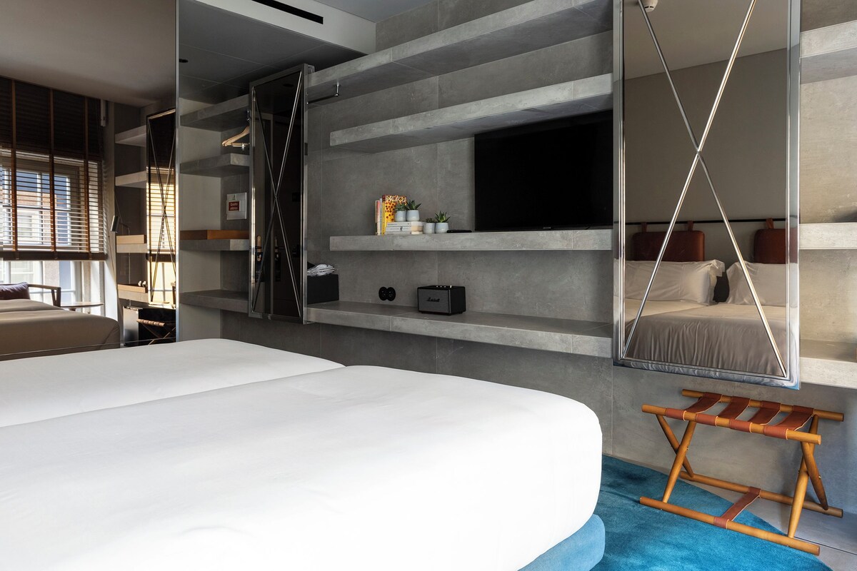 A modern hotel room with a large bed, a built-in TV, a seating area with a swing, and a small table with a lamp.