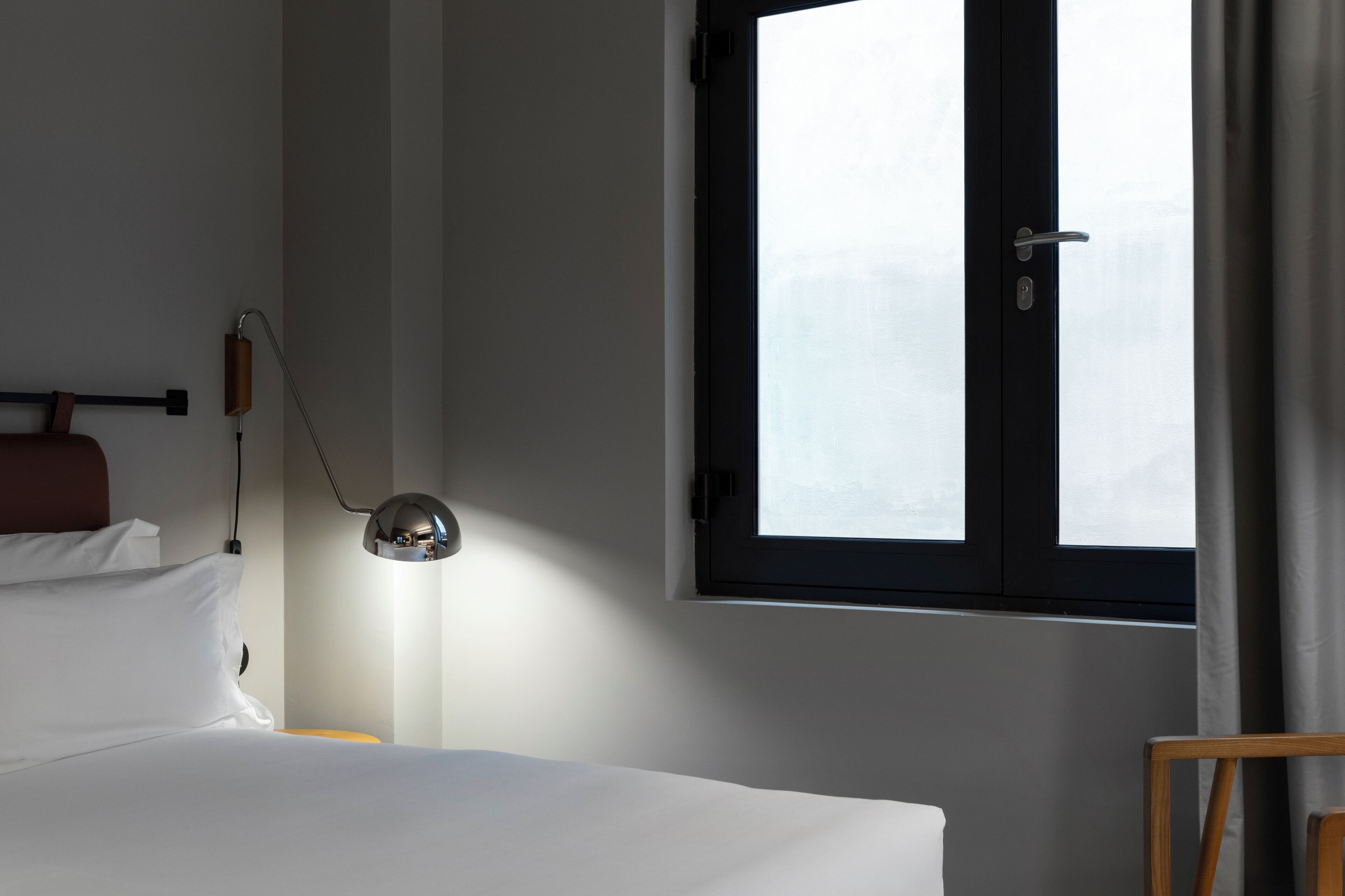 A hotel room with a bed, a desk lamp, a window with curtains, and a view of the outside.