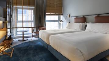 A hotel room with two beds, a desk, a chair, a lamp, and a view of the outside through large windows.